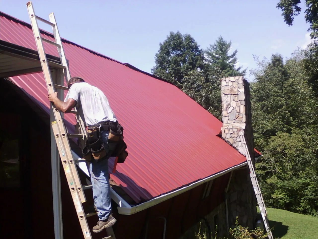 Roofer on a ladder beside a red metal roof and stone chimney — Soffit & Fascia Repair by Roofing Brothers in North Windham